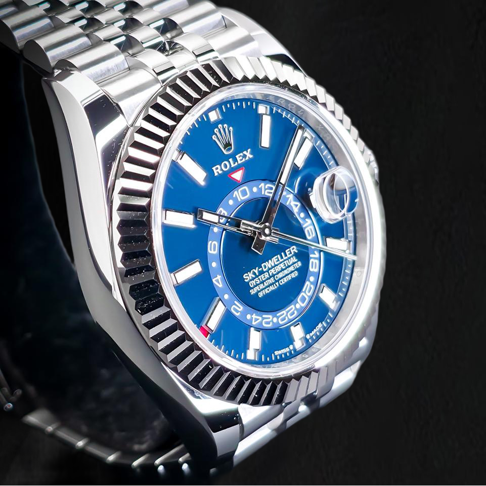 A Rolex Sky-Diver Date watch with a vibrant blue dial, silver stainless steel case and bracelet, and a fluted bezel, photographed against a dark textured background.